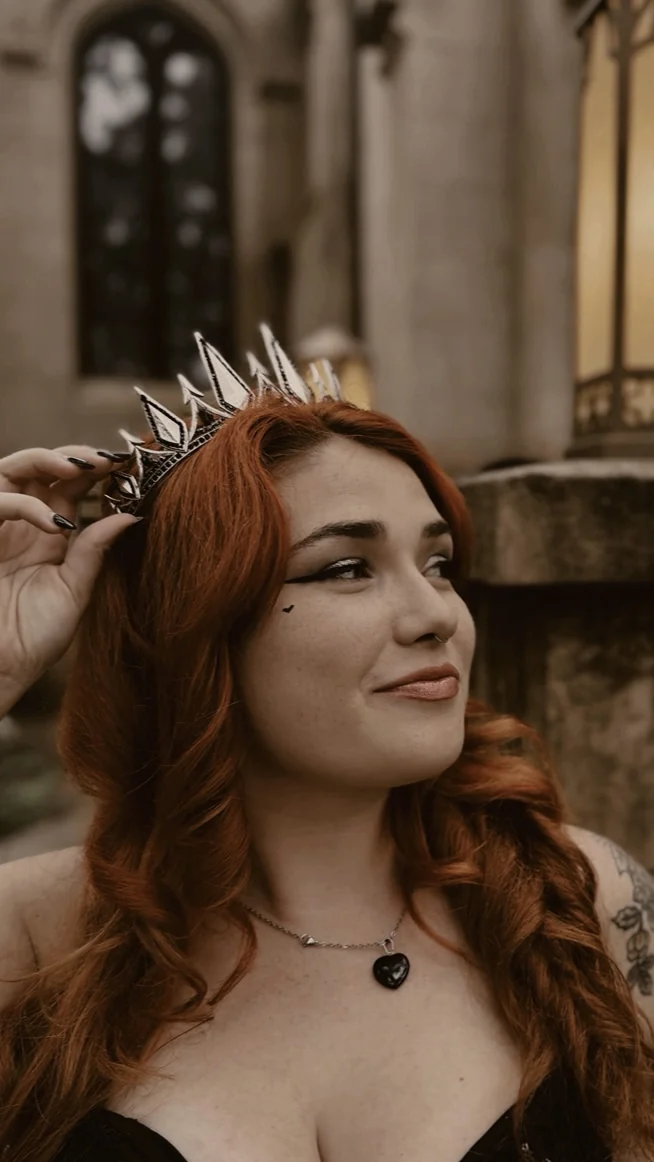 Pixie Rella in a silver crown, with long copper hair, in front of a gothic stone church window.